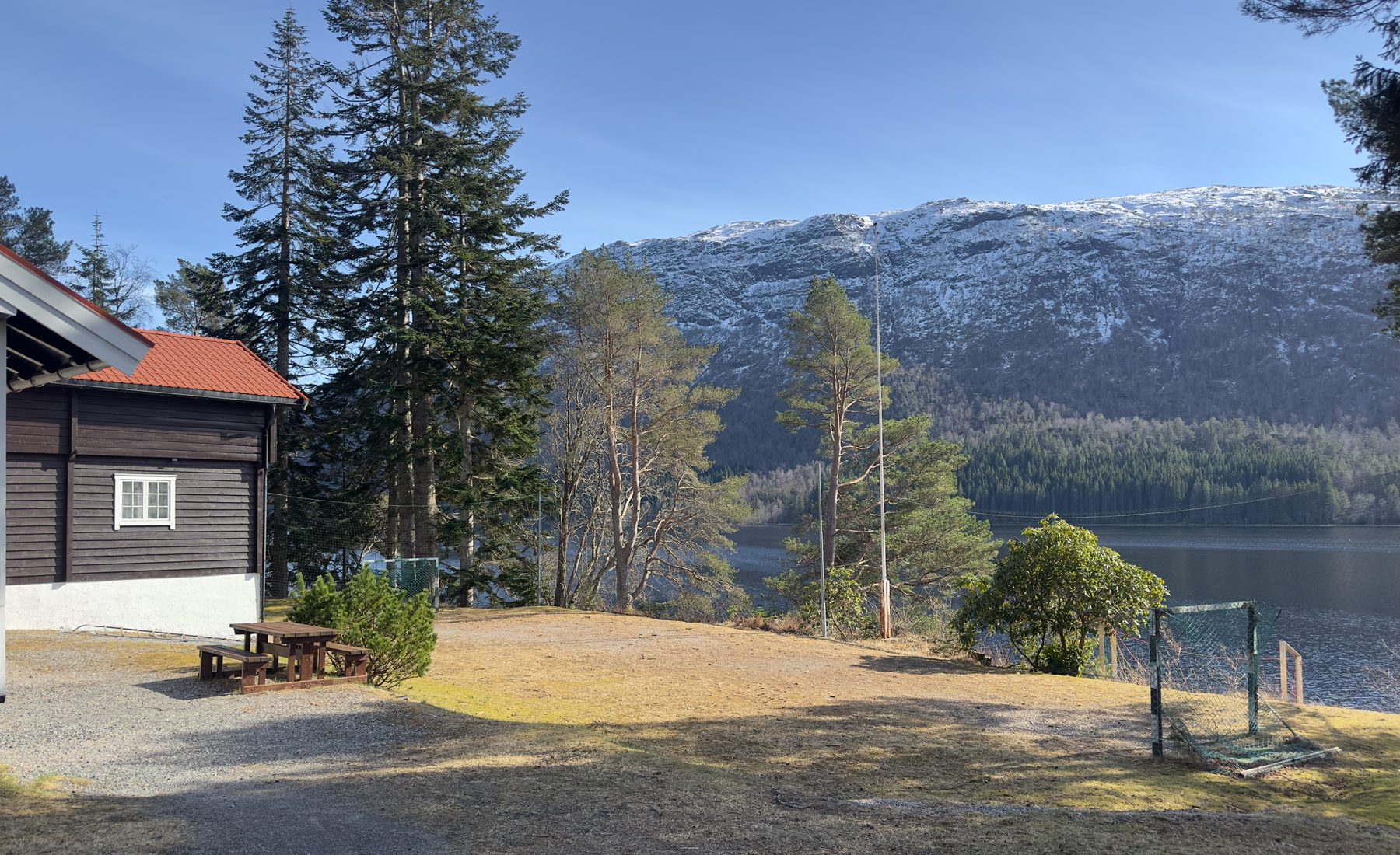 Mørk trehytte ved vatnet med fjell og skog i bakgrunnen
